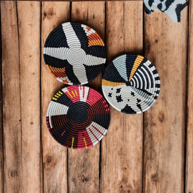 May include: Three woven decorative wall baskets in a variety of colors. The baskets are round with geometric patterns in black, white, orange, red, and yellow. The baskets are displayed on a wooden background.