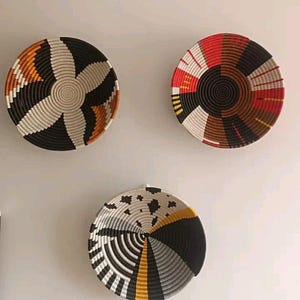 May include: Three decorative woven wall baskets in a variety of colors. The baskets are round with intricate patterns in black, white, orange, red, and brown. The baskets are mounted on a light gray wall.