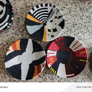 May include: A collection of woven decorative bowls in various colors. The bowls feature concentric circular patterns in black, white, red, yellow, and orange. The bowls are arranged on a textured surface.