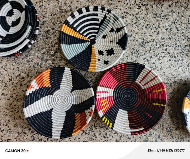 May include: A collection of woven decorative bowls in various colors. The bowls feature concentric circular patterns in black, white, red, yellow, and orange. The bowls are arranged on a textured surface.