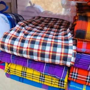 May include: A selection of folded plaid fabrics in various colours. The top fabric features a red, white, and black check pattern. Beneath it are purple, yellow, and blue plaid textiles, neatly stacked to display the different designs.