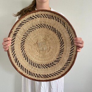May include: A large, round woven basket in natural beige and brown tones. The basket has a spiral design with alternating beige and dark brown bands. The rim is trimmed with a dark brown material.