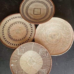 May include: Four round woven baskets in varying sizes and patterns. The baskets are primarily beige and brown, with intricate designs including geometric shapes and circular patterns. The rims are finished with a darker brown trim.