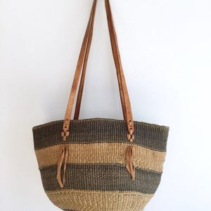 Może przedstawiać: Tkana torba typu tote z brązowymi i beżowymi poziomymi paskami. Torba ma dwa długie, brązowe skórzane paski i ozdobne skórzane frędzle. Torba wisi na białej ścianie.