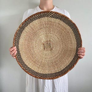 Pode incluir: Uma grande cesta redonda tecida com um centro bege natural e uma borda com padrão marrom escuro e preto. A cesta tem um design quadrado tecido no centro e é segurada por uma pessoa.