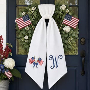 Könnte beinhalten: Ein weißer Türschal mit gestickten amerikanischen Flaggen und einem blauen Initial "W". Zwei kleine amerikanische Flaggen sind auf beiden Seiten eines grünen Kranzes platziert. Ein verzinkter Eimer mit Blumen und einer kleinen Flagge befindet sich links.