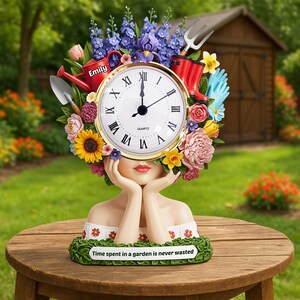 May include: A decorative clock featuring a woman's face with a floral headdress. The clock face has Roman numerals and gold trim. A red watering can with the name "Emily" is visible. The base reads "Time spent in a garden is never wasted."