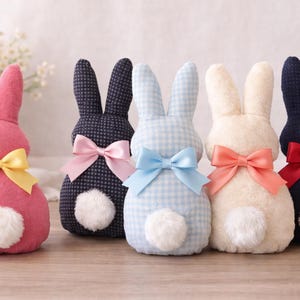 Puede incluir: Cinco figuras de conejito de peluche en rosa, azul marino, cuadros vichy azules, crema y azul marino con lazos. Cada conejito tiene una cola blanca y esponjosa. Los conejitos están colocados sobre una superficie marrón claro.