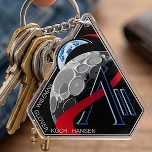 May include: A clear acrylic keychain with a space-themed design. The keychain features a black triangle with a stylized moon and Earth, a large letter "A", and the names GLOVER, WISEMAN, KOCH, and HANSEN. A silver chain holds the keychain and a set of keys.