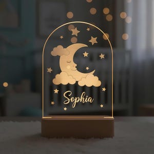 May include: A night light with a wooden base and a clear acrylic panel. The panel features a crescent moon with closed eyes, clouds, stars, and the name "Sophia" in script. The light has a warm glow.