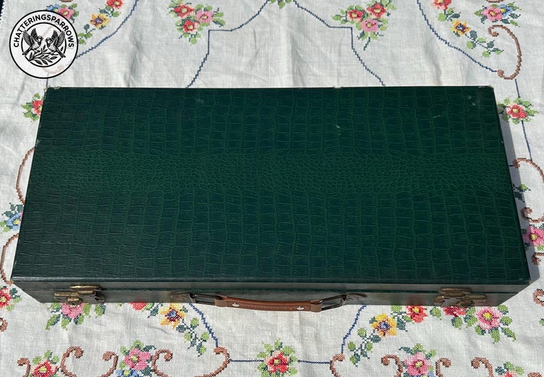 May include: A rectangular dark green storage case with a textured faux-alligator skin pattern. The case features a brown leather handle, two metallic latches on the front, and a circular logo in the top left corner with the text Chatteringsparrows.