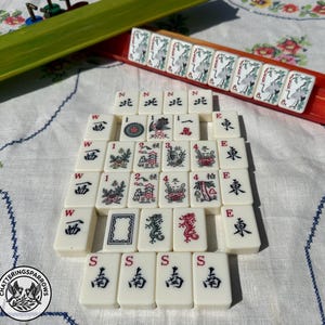 May include: A set of off-white Mahjong tiles featuring black and red ink illustrations of dragons, flowers, directional characters, and numbers. Some tiles are arranged in a rectangular grid, while others are placed in a red and green rack.