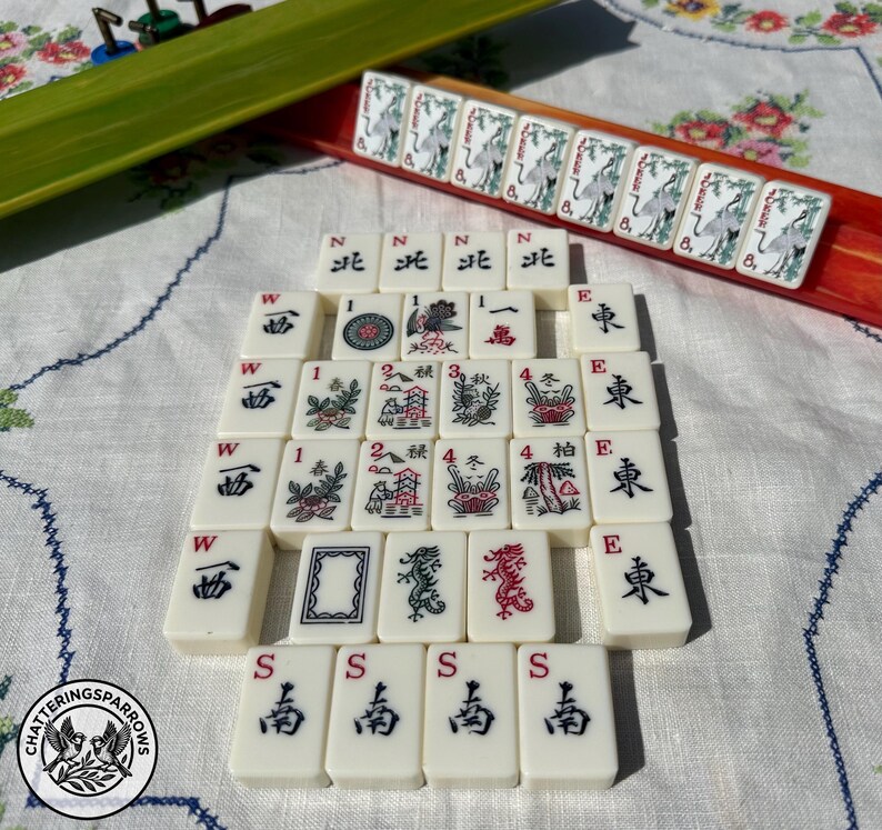 May include: A set of off-white Mahjong tiles featuring black and red ink illustrations of dragons, flowers, directional characters, and numbers. Some tiles are arranged in a rectangular grid, while others are placed in a red and green rack.