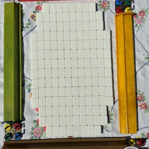 May include: A set of white mahjong tiles arranged in a rectangular grid, surrounded by four long, colored tile racks in red, green, yellow, and brown. Each rack features small, circular, multi-colored scoring markers at the ends.