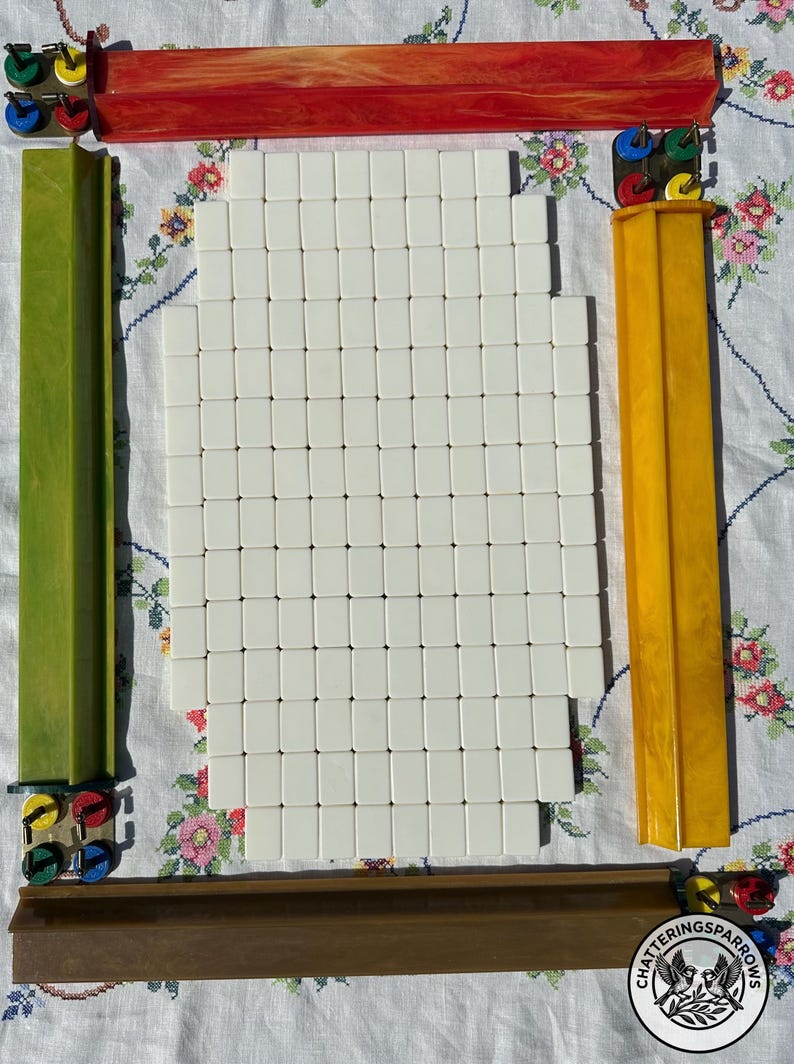May include: A set of white mahjong tiles arranged in a rectangular grid, surrounded by four long, colored tile racks in red, green, yellow, and brown. Each rack features small, circular, multi-colored scoring markers at the ends.