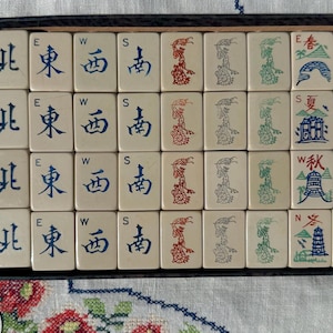May include: A set of off-white Mahjong tiles arranged in a rectangular box. The tiles feature blue Chinese characters for North, East, West, and South, red and green dragon illustrations, and seasonal imagery with red, blue, and green ink details.