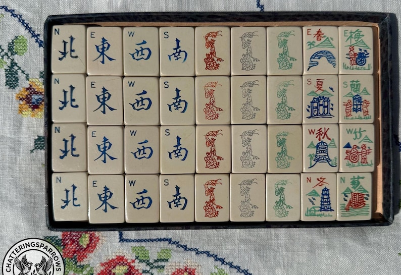 May include: A set of off-white Mahjong tiles arranged in a rectangular box. The tiles feature blue Chinese characters for North, East, West, and South, red and green dragon illustrations, and seasonal imagery with red, blue, and green ink details.