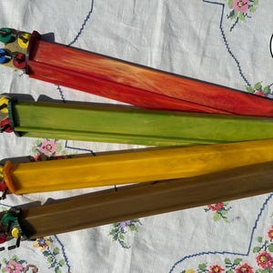 May include: Four long, rectangular knitting needles with marbled finishes in red, green, yellow, and brown. Each needle features a distinct end cap with small, colorful circular accents in green, yellow, blue, and red, arranged in a cluster.