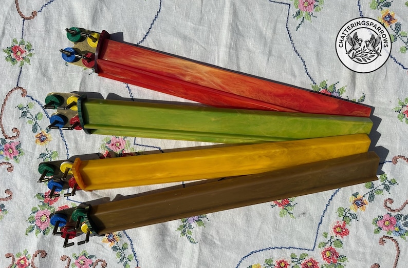 May include: Four long, rectangular knitting needles with marbled finishes in red, green, yellow, and brown. Each needle features a distinct end cap with small, colorful circular accents in green, yellow, blue, and red, arranged in a cluster.