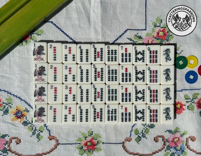 May include: A set of off-white Mahjong tiles arranged in four rows, featuring black and red ink designs including numbers one through nine, bird illustrations, and dragon symbols. A circular logo with two birds and the text CHATTERINGSPARROWS is visible.