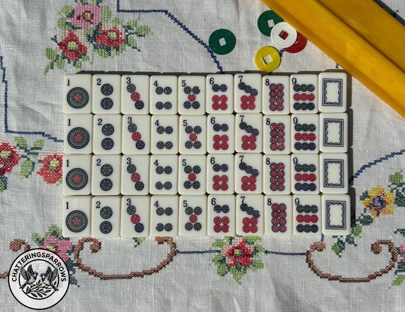 May include: A set of off-white Mahjong tiles arranged in a grid, featuring numbers one through nine in red and black ink, alongside blank tiles. A yellow plastic rack and several small, circular, colored plastic tokens are positioned nearby.