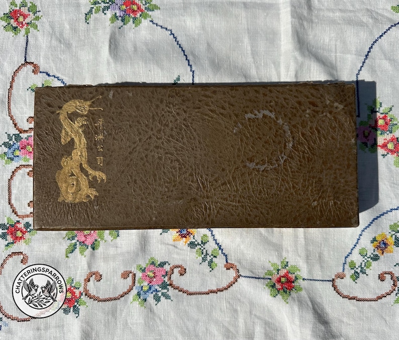 May include: A rectangular brown box with a textured surface featuring a gold-coloured dragon illustration on the left side. The box includes vertical gold-coloured Japanese characters next to the dragon and a circular mark on the right side.
