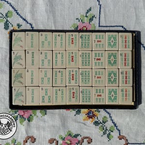 May include: A rectangular black tray containing thirty-two off-white mahjong tiles arranged in four rows of eight. Each tile features green and red engraved numerical markings and symbols, including birds, bamboo patterns, and Arabic numerals one through nine.