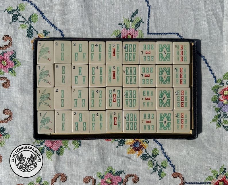 May include: A rectangular black tray containing thirty-two off-white mahjong tiles arranged in four rows of eight. Each tile features green and red engraved numerical markings and symbols, including birds, bamboo patterns, and Arabic numerals one through nine.