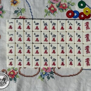 May include: A set of white Mahjong tiles arranged in four rows, featuring black numbers one through nine, red Chinese characters, and red dragon symbols. Five small, round, colored plastic counting tokens are placed in the upper right corner.