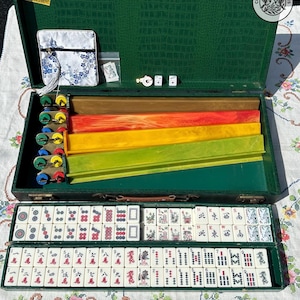 May include: A Mahjong set in a green textured case containing four colorful tile racks, white tiles with red, black, and green symbols, two dice, and a small white fabric pouch with a blue floral pattern. The tiles feature Chinese characters and directional markers.