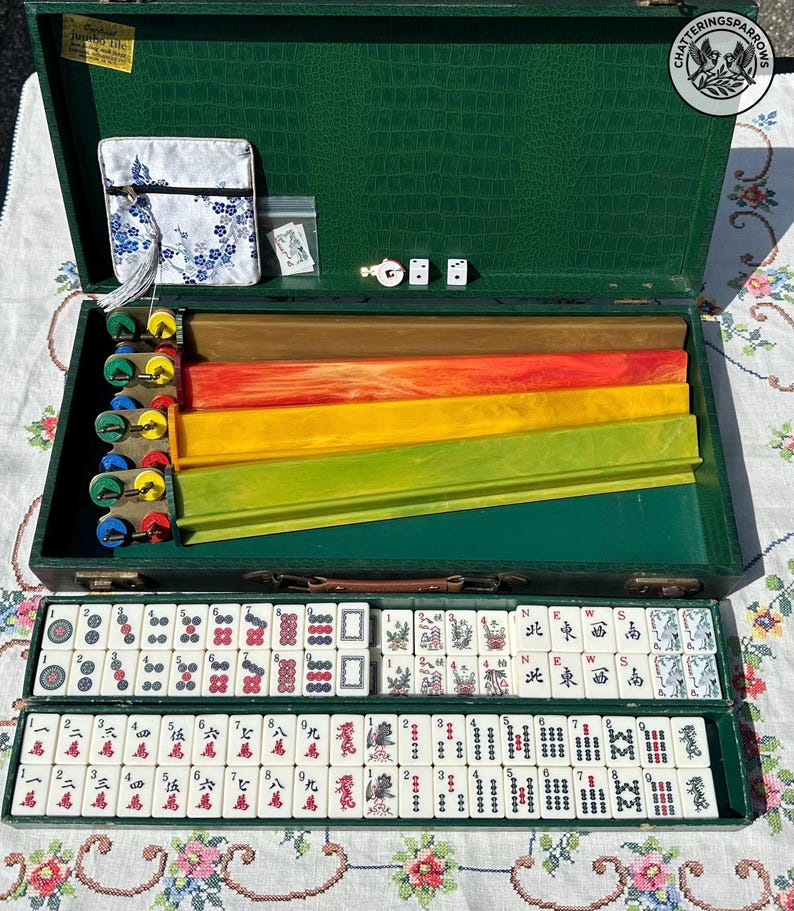May include: A Mahjong set in a green textured case containing four colorful tile racks, white tiles with red, black, and green symbols, two dice, and a small white fabric pouch with a blue floral pattern. The tiles feature Chinese characters and directional markers.