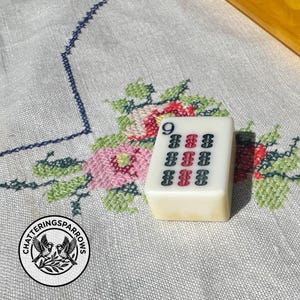 May include: A rectangular off-white Mahjong tile featuring the number 9 in the top left corner and a grid of nine vertical symbols in black and red ink. The tile has a smooth, polished surface and is positioned upright on a textured fabric surface.