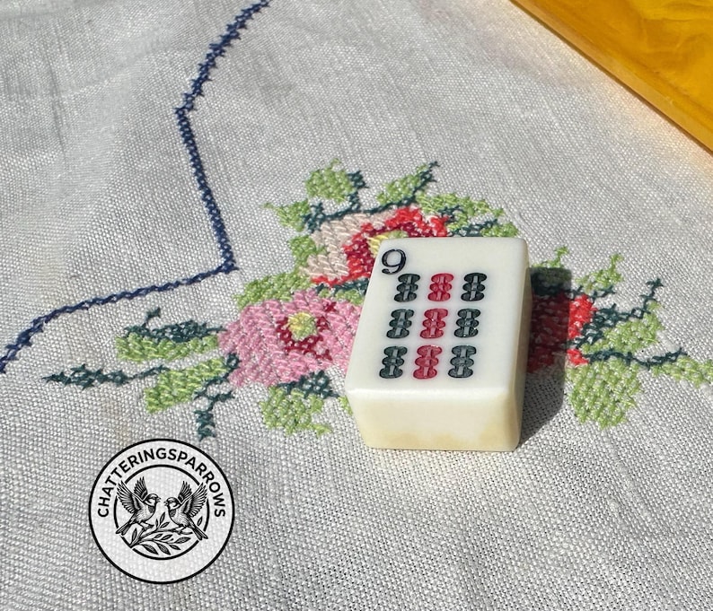 May include: A rectangular off-white Mahjong tile featuring the number 9 in the top left corner and a grid of nine vertical symbols in black and red ink. The tile has a smooth, polished surface and is positioned upright on a textured fabric surface.
