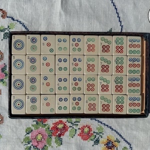 May include: A rectangular box containing thirty-two off-white mahjong tiles arranged in four rows of eight. Each tile features a numerical digit from one to nine, accompanied by circular symbols in red, blue, and green ink, set within a dark frame.