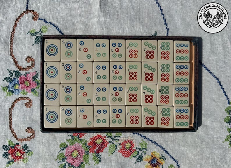 May include: A rectangular box containing thirty-two off-white mahjong tiles arranged in four rows of eight. Each tile features a numerical digit from one to nine, accompanied by circular symbols in red, blue, and green ink, set within a dark frame.