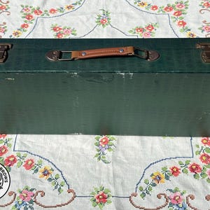 May include: A rectangular dark green storage case with a textured surface, featuring a brown leather-like handle and two metallic latches on the top. The case has visible wear along the edges and corners, with a solid construction and a flat lid.
