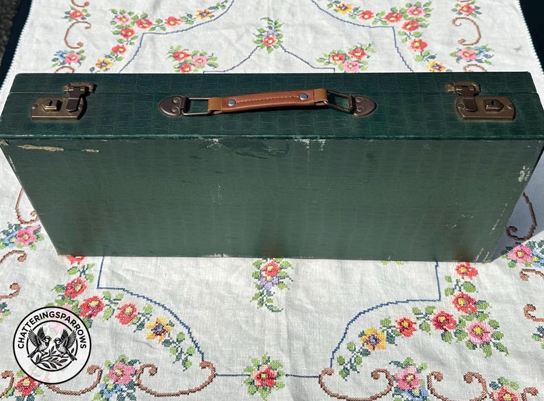 May include: A rectangular dark green storage case with a textured surface, featuring a brown leather-like handle and two metallic latches on the top. The case has visible wear along the edges and corners, with a solid construction and a flat lid.