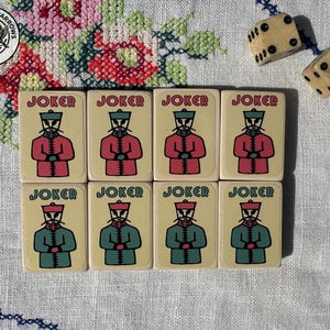 May include: Eight rectangular game tiles arranged in two rows of four, each featuring the word JOKER and a stylised illustration of a figure in a uniform. The top row figures wear red coats, while the bottom row figures wear green coats. Two dice are nearby.