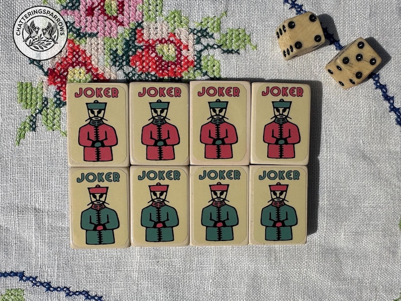 May include: Eight rectangular game tiles arranged in two rows of four, each featuring the word JOKER and a stylised illustration of a figure in a uniform. The top row figures wear red coats, while the bottom row figures wear green coats. Two dice are nearby.