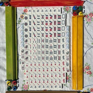 May include: A complete set of white Mahjong tiles featuring red, green, and black ink characters, dots, and bamboo symbols, arranged in a rectangular grid. The set includes four long, colored plastic tile racks in red, green, yellow, and brown.