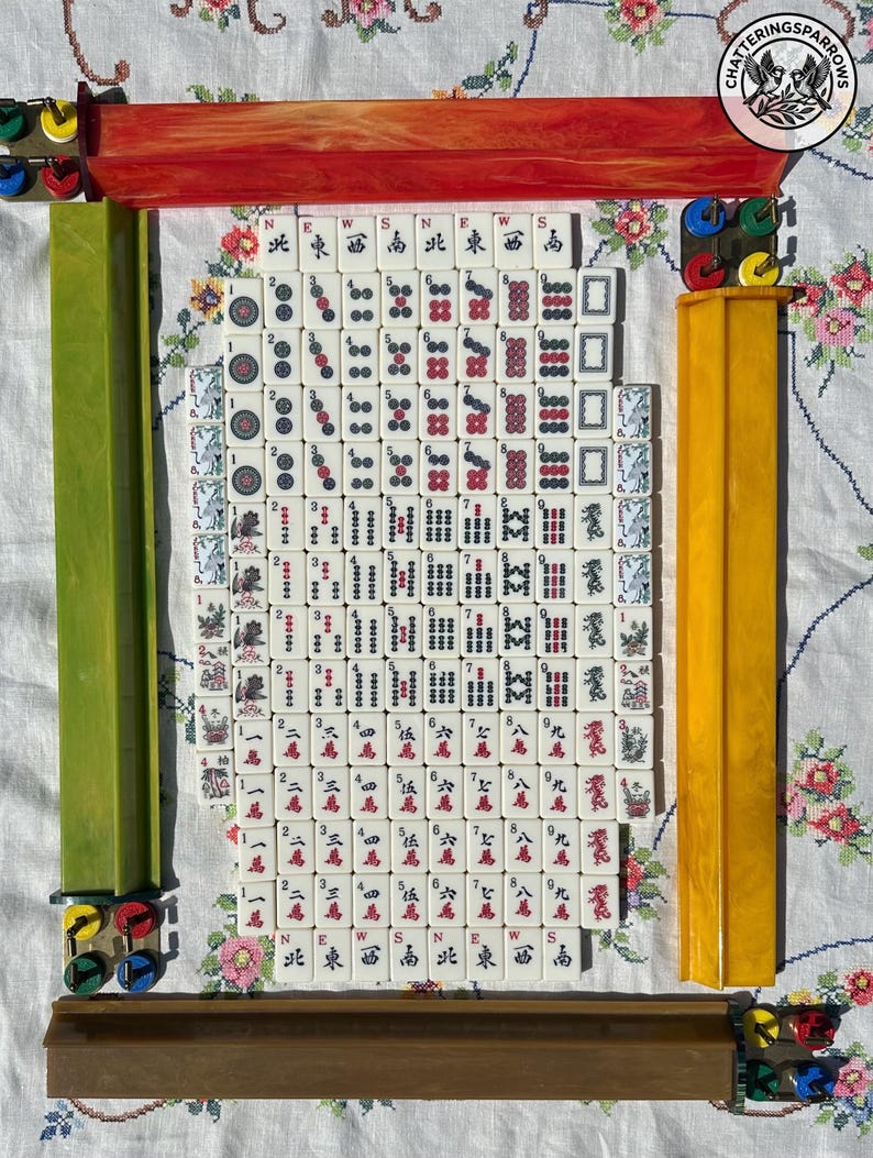 May include: A complete set of white Mahjong tiles featuring red, green, and black ink characters, dots, and bamboo symbols, arranged in a rectangular grid. The set includes four long, colored plastic tile racks in red, green, yellow, and brown.