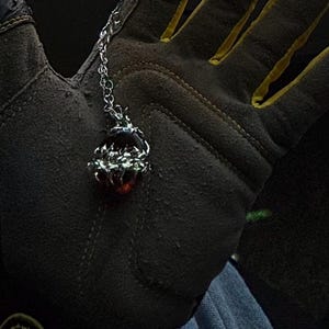 May include: A silver-toned pendant necklace featuring a dark red gemstone. The pendant has a detailed, ornate design. The necklace is displayed against a dark background, with a work glove in the background.