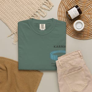 May include: A teal Comfort Colors t-shirt with the word "Karma" and a blue graphic design. The phrase "Every come" is printed below the graphic. The shirt is displayed with beige pants, a woven mat, and a candle.