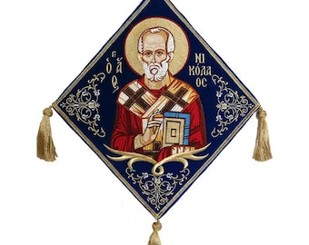 May include: A diamond-shaped religious icon featuring an embroidered image of a saint with a halo. The icon has a dark blue background with gold accents and tassels. The saint is depicted in red and gold robes, holding a book.