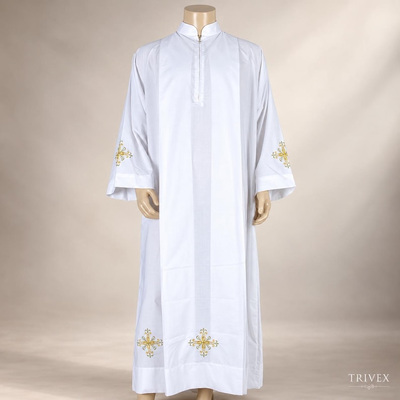 May include: A white liturgical alb featuring a mandarin collar and a front zipper closure. The garment is decorated with gold and silver embroidered cross motifs positioned on the lower hem and on the outer side of each wide sleeve cuff.