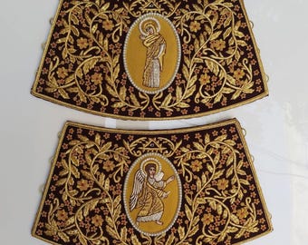 May include: Two ornate, embroidered religious vestments. Each features a dark brown background with gold floral patterns and a central oval with a religious figure. The edges are trimmed with gold and red accents.
