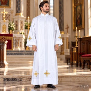 May include: A white liturgical alb featuring a high collar, long sleeves, and gold embroidered cross motifs on the cuffs and lower hem. The garment has a central vertical panel design and is worn with black dress shoes in a church setting.
