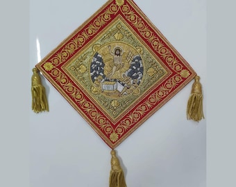 May include: A diamond-shaped religious tapestry with a red border and gold embroidery. The center features a detailed illustration with a figure in a golden halo. Gold tassels hang from the corners, adding a decorative touch to this religious item.
