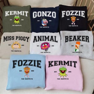 May include: A selection of sweatshirts showcasing Muppets characters. Each sweatshirt has a character's name in bold, an image of the character, and the text "EST. 1955 THE MUPPETS". The sweatshirts come in colours such as green, navy, black, beige, white, grey, and light blue.