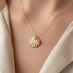 May include: A gold necklace featuring a delicate chain and a pendant shaped like a scalloped seashell. The center of the seashell pendant is accented with a small, round white pearl. The necklace rests against a light beige collared shirt.